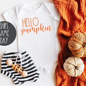 May include: White baby onesie with the text "HELLO pumpkin" in orange. The onesie is lying on a white surface with a pair of black and white striped baby pants and a wooden rattle. A brown fabric with two decorative pumpkins is in the background.