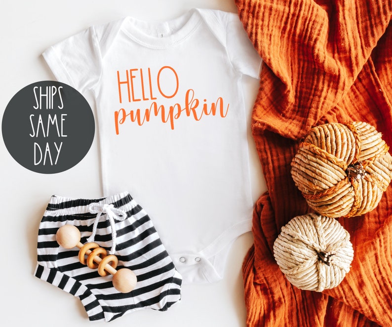 May include: White baby onesie with the text "HELLO pumpkin" in orange. The onesie is lying on a white surface with a pair of black and white striped baby pants and a wooden rattle. A brown fabric with two decorative pumpkins is in the background.