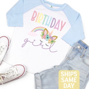 K&ouml;nnte beinhalten: Ein wei&szlig;es und hellblaues Baseball-T-Shirt mit einem Regenbogen- und Einhorn-Design. Das Shirt tr&auml;gt die Aufschrift "Birthday girl" in lila Schrift.