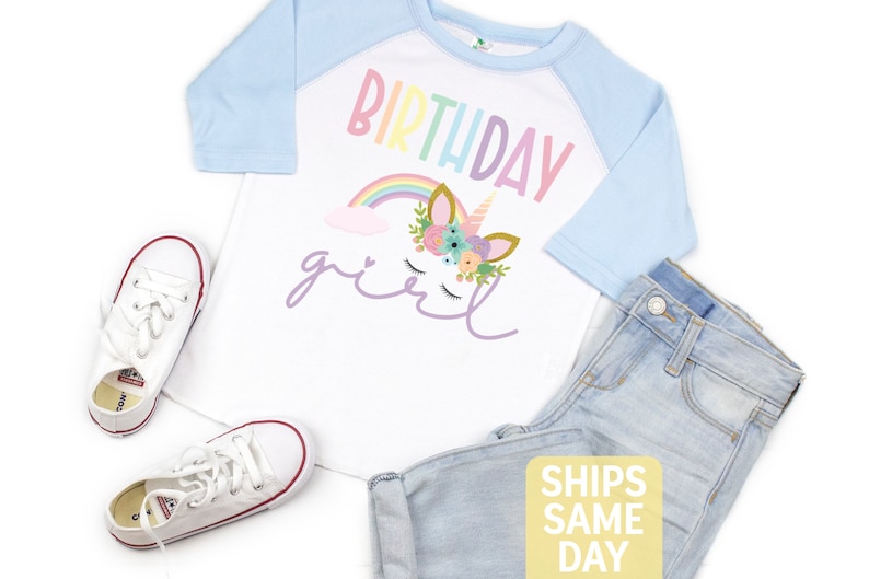 K&ouml;nnte beinhalten: Ein wei&szlig;es und hellblaues Baseball-T-Shirt mit einem Regenbogen- und Einhorn-Design. Das Shirt tr&auml;gt die Aufschrift "Birthday girl" in lila Schrift.