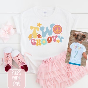 May include: A white t-shirt with the words "TWO GROOVY" in colorful retro font, a peace sign, and flower graphics. The shirt is paired with a pink tutu skirt and pink sneakers. A photo shows the back of a shirt with the name "SOPHIA" and the number "2".