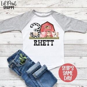 May include: A white and gray raglan t-shirt with a red barn and farm animals. The number 3 is in black and white polka dots. The name Rhett is printed in black below the barn.