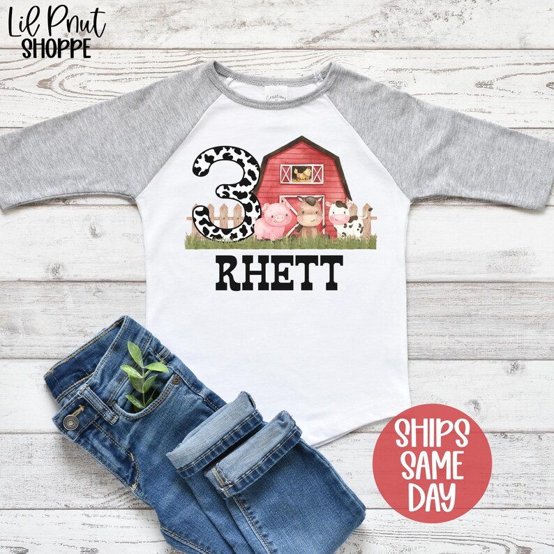 May include: A white and gray raglan t-shirt with a red barn and farm animals. The number 3 is in black and white polka dots. The name Rhett is printed in black below the barn.