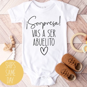 May include: White baby onesie with black text that reads "¡Sorpresa! Vas a ser abuelito" with a heart underneath. The onesie is on a wooden background with brown baby shoes, a wooden teether, and a beige and brown baby toy.