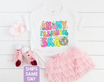 May include: White t-shirt with a colorful graphic that says "In My Preschool Era" with a pencil, apple, and disco ball. The shirt is paired with a pink tutu and pink sneakers.