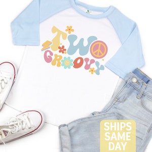 May include: A white and light blue raglan shirt with the text "Two Groovy" in colorful retro font, paired with light wash denim jeans and white canvas sneakers. The shirt features a peace sign and flower graphics.