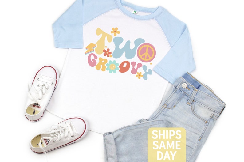 May include: A white and light blue raglan shirt with the text "Two Groovy" in colorful retro font, paired with light wash denim jeans and white canvas sneakers. The shirt features a peace sign and flower graphics.