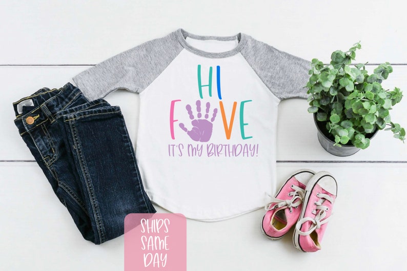 K&ouml;nnte beinhalten: Ein wei&szlig;es und graues Baseball-T-Shirt mit dem Text "Hi Five It's My Birthday!" in bunten Buchstaben mit einem lila Handabdruck in der Mitte des Wortes "Five".