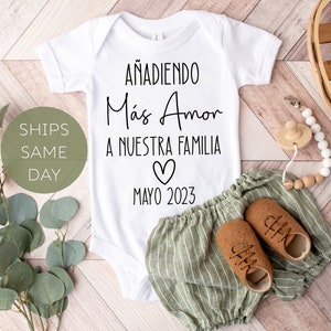 May include: White baby onesie with black text that reads "Añadiendo Más Amor A Nuestra Familia" with a heart and the date "Mayo 2023".