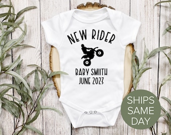 May include: White baby onesie with black text that reads "NEW RIDER" with a silhouette of a person riding an ATV. The text "BABY SMITH JUNE 2023" is below the ATV.