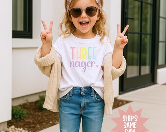 May include: A young child wearing a white t-shirt with the words "THREE nager" in pastel colors. The child is also wearing sunglasses, a headband, and blue jeans. The child is smiling and making a peace sign with their fingers.