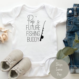 May include: White baby onesie with black text that reads "Pop's Future Fishing Buddy" and a black silhouette of a fishing rod.  The onesie is laying on a white surface with a pair of gray baby shoes, a wooden baby toy, and a pair of blue jeans.