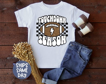 May include: White t-shirt with a brown football graphic and the text "Touchdown Season" in white letters with a checkered background.