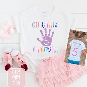 Pu&ograve; includere: Maglietta bianca con il testo "Officially a Handful" e un'impronta di mano viola con il numero 5 al suo interno. La parte posteriore della maglietta ha il nome "Chloe" e il numero 5 in colori arcobaleno.