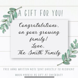 May include: A white card with a handwritten message that reads "A Gift For You! Congratulations, on your growing family! Love, The Smith Family". The card is surrounded by green leafy branches.