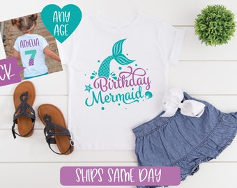 May include: White t-shirt with a teal mermaid tail and the text "Birthday Mermaid" in purple. The shirt is on a white surface with a pair of black sandals and a blue denim skirt with a white bow.