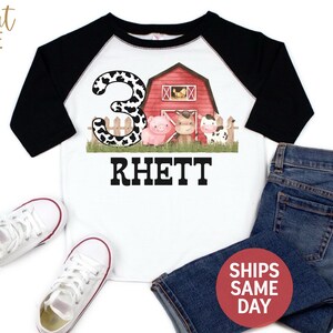 May include: A white and black raglan t-shirt with a red barn, a pig, a cow, and a horse. The number 3 is in the background with a cow print design. The name Rhett is printed below the farm scene.