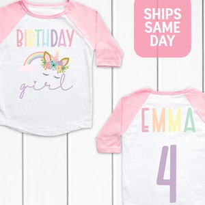 K&ouml;nnte beinhalten: Wei&szlig;es und rosafarbenes Baseball-Shirt mit einem Regenbogen-Einhorn und dem Text "Birthday girl". Auf der R&uuml;ckseite des Shirts steht der Name "Emma" in Regenbogenfarben und die Zahl "4" in Lila.