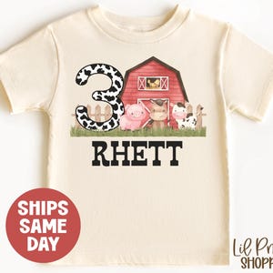 May include: A white t-shirt with a red barn, a cow, a pig, and a number 3 with black and white cow spots. The text "Rhett" is printed below the barn.