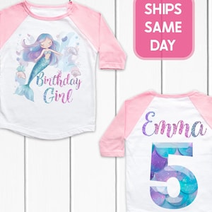 May include: White and pink raglan-style birthday shirt. The front features a mermaid illustration and the text "Birthday Girl". The back has the name "Emma" and the number "5" in a mermaid scale design.