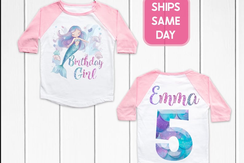 May include: White and pink raglan-style birthday shirt. The front features a mermaid illustration and the text "Birthday Girl". The back has the name "Emma" and the number "5" in a mermaid scale design.