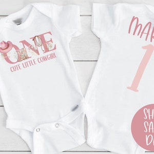 May include: White onesie with pink text that says "ONE" and "Cute Little Cowgirl". The onesie has a pink cowboy hat and boots graphic. The back of the onesie has the name "MAREN" and the number "1" in pink.