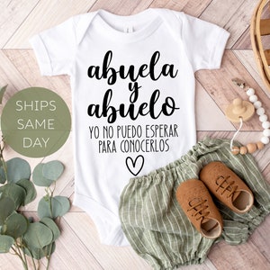 May include: White baby onesie with the text "abuela y abuelo yo no puedo esperar para conocerlos" in black lettering with a small heart at the bottom. The onesie is laying on a wooden surface with green and white striped pants, brown shoes, and a wooden and white bead necklace.