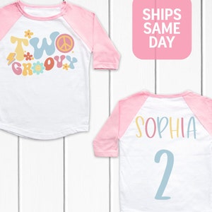 May include: White and pink raglan style t-shirt with the words "TWO GROOVY" in colorful, retro font on the front. The back of the shirt has the name "SOPHIA" and the number "2" in a similar style.
