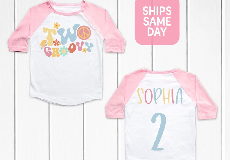 May include: White and pink raglan style t-shirt with the words "TWO GROOVY" in colorful, retro font on the front. The back of the shirt has the name "SOPHIA" and the number "2" in a similar style.