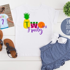May include: White t-shirt with a colorful graphic that says "Two Fruity" with a pineapple, watermelon, and orange. The shirt is on a white surface with a pair of black sandals, a denim skirt, and a small green plant.