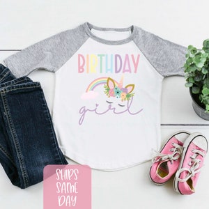 K&ouml;nnte beinhalten: Wei&szlig;es Langarmshirt mit grauem Baseball-&Auml;rmeldesign. Das Shirt hat einen Regenbogen, eine Wolke und ein Einhorn mit Blumen auf dem Kopf. Der Text "Birthday girl" steht auf dem Shirt.