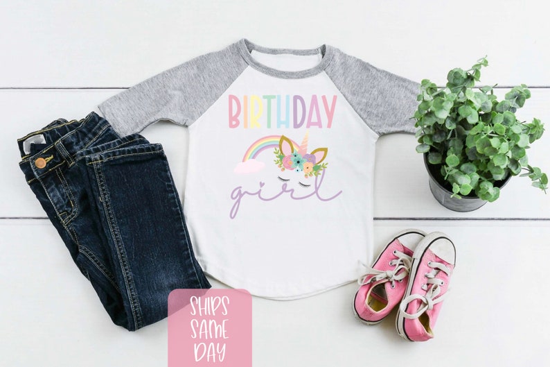K&ouml;nnte beinhalten: Wei&szlig;es Langarmshirt mit grauem Baseball-&Auml;rmeldesign. Das Shirt hat einen Regenbogen, eine Wolke und ein Einhorn mit Blumen auf dem Kopf. Der Text "Birthday girl" steht auf dem Shirt.