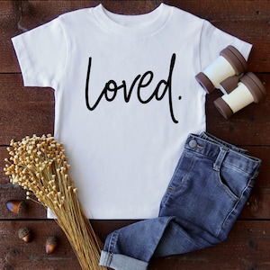 K&ouml;nnte beinhalten: Wei&szlig;es T-Shirt mit dem Wort "loved" in schwarzer Schrift auf einem Holzgrund.