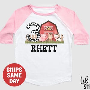May include: A pink and white baseball style t-shirt with a cow print number 3, a red barn, and a pig, cow, and horse. The text "Rhett" is below the image.