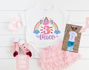 May include: White t-shirt with a rainbow, unicorn, and the number 3 with the word "three" underneath. The shirt is shown on a white background with a pink tutu, pink shoes, and a pink bow. The back of the shirt is shown in a separate image with the name "Luna" and the number 3.
