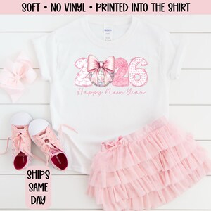 May include: White t-shirt with a pink bow and the numbers "2026" in a pink watercolor design, with "Happy New Year" text. The shirt is paired with a pink tutu skirt and pink shoes. A pink bow is also present.