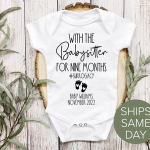 May include: White baby onesie with black text that reads "With the Babysitter for Nine Months #Surrogacy" and "Baby Williams November 2022".  The onesie has a pair of black footprints with hearts on them.
