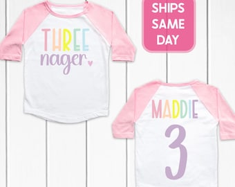 May include: White and pink raglan-style toddler shirt. The front displays the text "THREE nager" in pastel colors. The back of the shirt has the name "MADDIE" and the number "3" in pastel colors. The shirt has three-quarter sleeves.