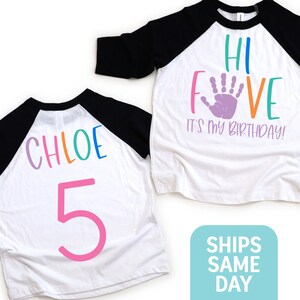 K&ouml;nnte beinhalten: Zwei wei&szlig;e Baseball-Shirts mit schwarzen &Auml;rmeln. Ein Shirt sagt "Hi Five It's My Birthday!" mit einem lila Handabdruck. Das andere Shirt sagt "Chloe 5" mit Regenbogenbuchstaben und einer pinken Zahl 5.