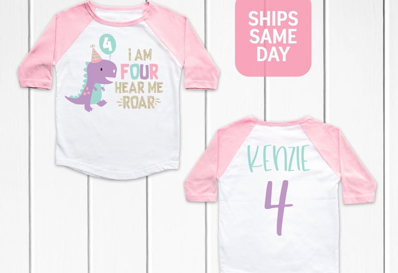 May include: A white and pink baseball style shirt with a purple dinosaur graphic and the text "I am four hear me roar" on the front. The back of the shirt has the name "Kenzie" and the number "4" in purple.