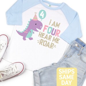 May include: A white and light blue raglan t-shirt with a purple dinosaur wearing a party hat and holding a balloon with the number 4 on it. The shirt also has the text "I am Four Hear Me Roar" in rainbow colors with gold glitter.