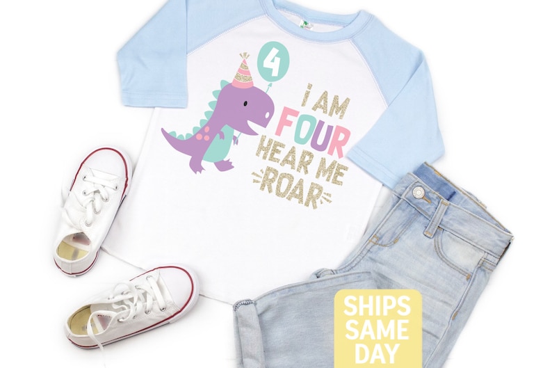 May include: A white and light blue raglan t-shirt with a purple dinosaur wearing a party hat and holding a balloon with the number 4 on it. The shirt also has the text "I am Four Hear Me Roar" in rainbow colors with gold glitter.