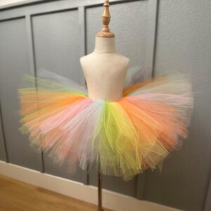 May include: A rainbow-colored tutu skirt displayed on a mannequin. The skirt features layers of tulle in shades of pink, yellow, orange, and white. The tutu is designed for children's wear and is displayed against a gray wall.