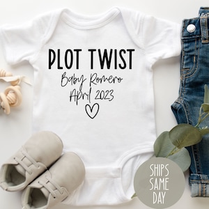 May include: White baby onesie with black text that reads "Plot Twist Baby Romero April 2023" with a small heart symbol.