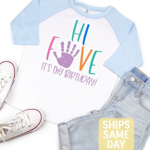 K&ouml;nnte beinhalten: Wei&szlig;es T-Shirt mit einem blau-wei&szlig;en Baseball-&Auml;rmel. Das Shirt hat einen lila Handabdruck und den Text "Hi Five It's My Birthday!" in Regenbogenfarben.