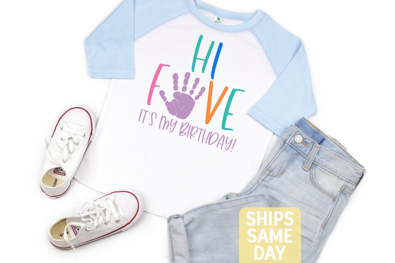 K&ouml;nnte beinhalten: Wei&szlig;es T-Shirt mit einem blau-wei&szlig;en Baseball-&Auml;rmel. Das Shirt hat einen lila Handabdruck und den Text "Hi Five It's My Birthday!" in Regenbogenfarben.