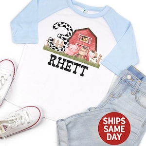 May include: A white and light blue baseball tee shirt with a farm animal graphic and the number 3. The shirt says "Rhett" on the front.