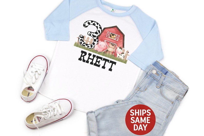 May include: A white and light blue baseball tee shirt with a farm animal graphic and the number 3. The shirt says "Rhett" on the front.