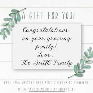 May include: A white card with the text "A GIFT FOR YOU!" and "Congratulations, on your growing family! Love. The Smith Family." The card is decorated with green leaf sprigs and is on a white wood-paneled background.