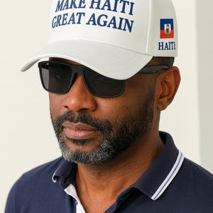 May include: White baseball cap with the text "MAKE HAITI GREAT AGAIN" in blue. The cap also features the Haitian flag and the word "HAITI". The person in the image is wearing black sunglasses and a navy blue polo shirt.